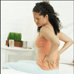 Woman sitting on the edge of her bed holding her lower back, experiencing morning back stiffness and pain.